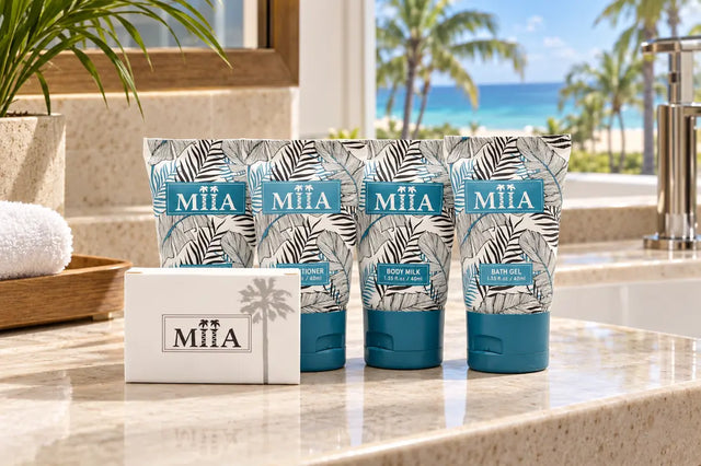 Four MiiA body care tubes and a matching box arranged on a bathroom counter with a tropical beach view in the background. The packaging features a gray palm leaf pattern with teal blue labels and caps.