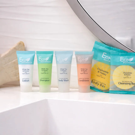 A set of Eco Botanics body care travel-size tubes and cleansing bar packets arranged on a white bathroom counter in front of a mirror, with a ceramic vessel on the left.