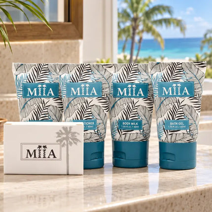 Four MiiA body care tubes and a matching box arranged on a bathroom counter with a tropical beach view in the background. The packaging features a gray palm leaf pattern with teal blue labels and caps.