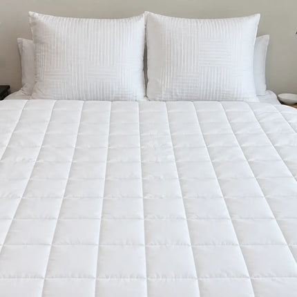 A white quilted bedspread with matching white pillows on a bed, shown in a styled bedroom setting with bedside tables and lamps.
