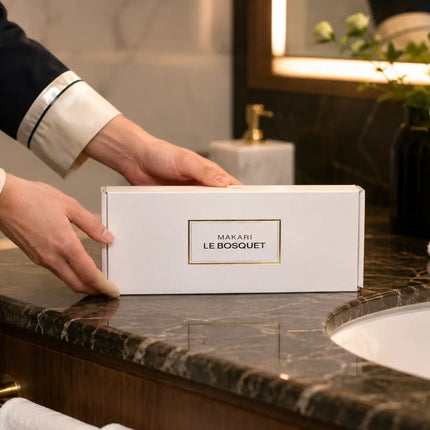 A person in a dark navy and cream uniform places a white rectangular box labeled 'MAJORET LE BOSQUET' on a dark marble bathroom counter next to a sink.